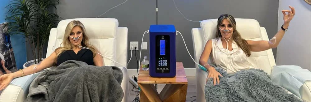 Two patients reclining side by side, each breathing hydrogen through a nasal cannula from a HydroHeal unit between them.